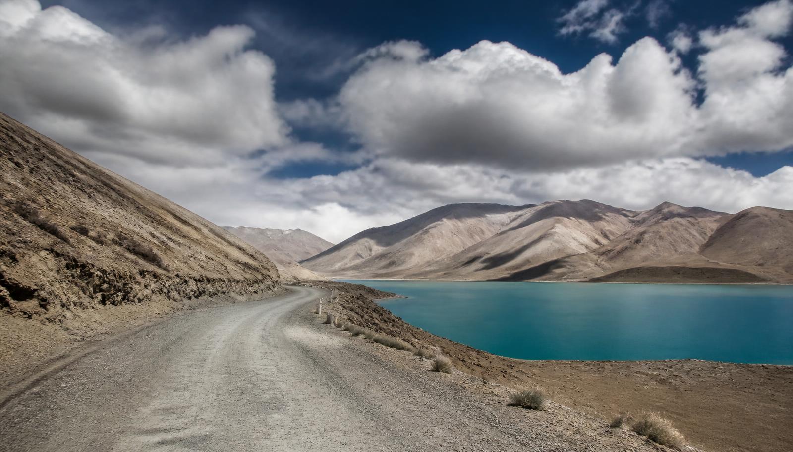 Empty gravel section of the Pamir Highway beside a turquoise lake in Tajikistan